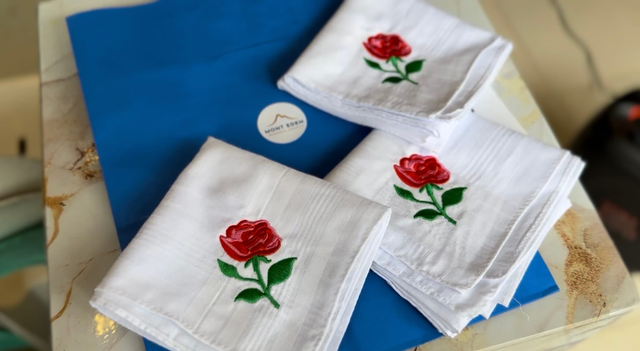 Three white handkerchiefs embroidered with red roses resting on a blue box, serving as commemorative keepsakes.