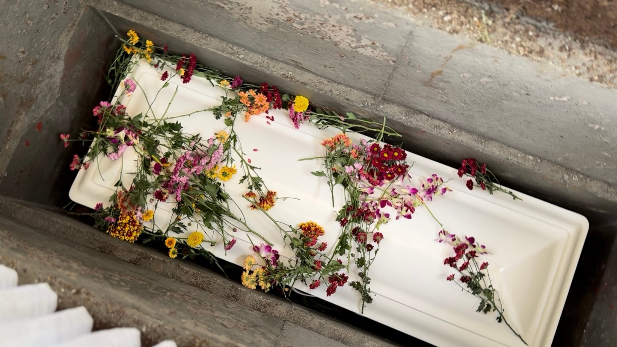 A white coffin adorned with loose, informally scattered fresh flowers in a concrete grave, showing a natural, personal floral farewell.
