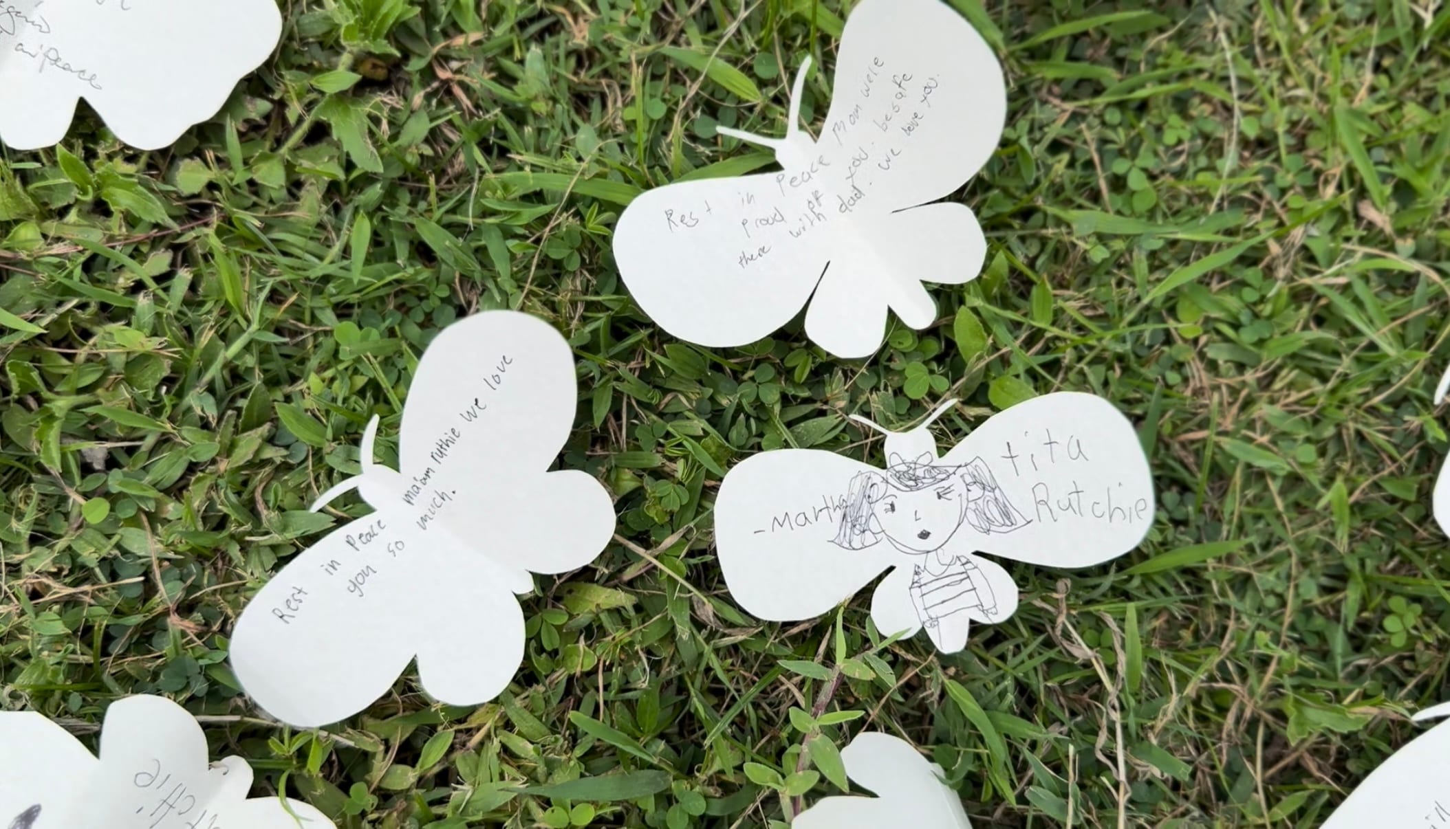 White paper butterfly cut-outs with handwritten messages and drawings placed on green grass as outdoor memorial tributes.