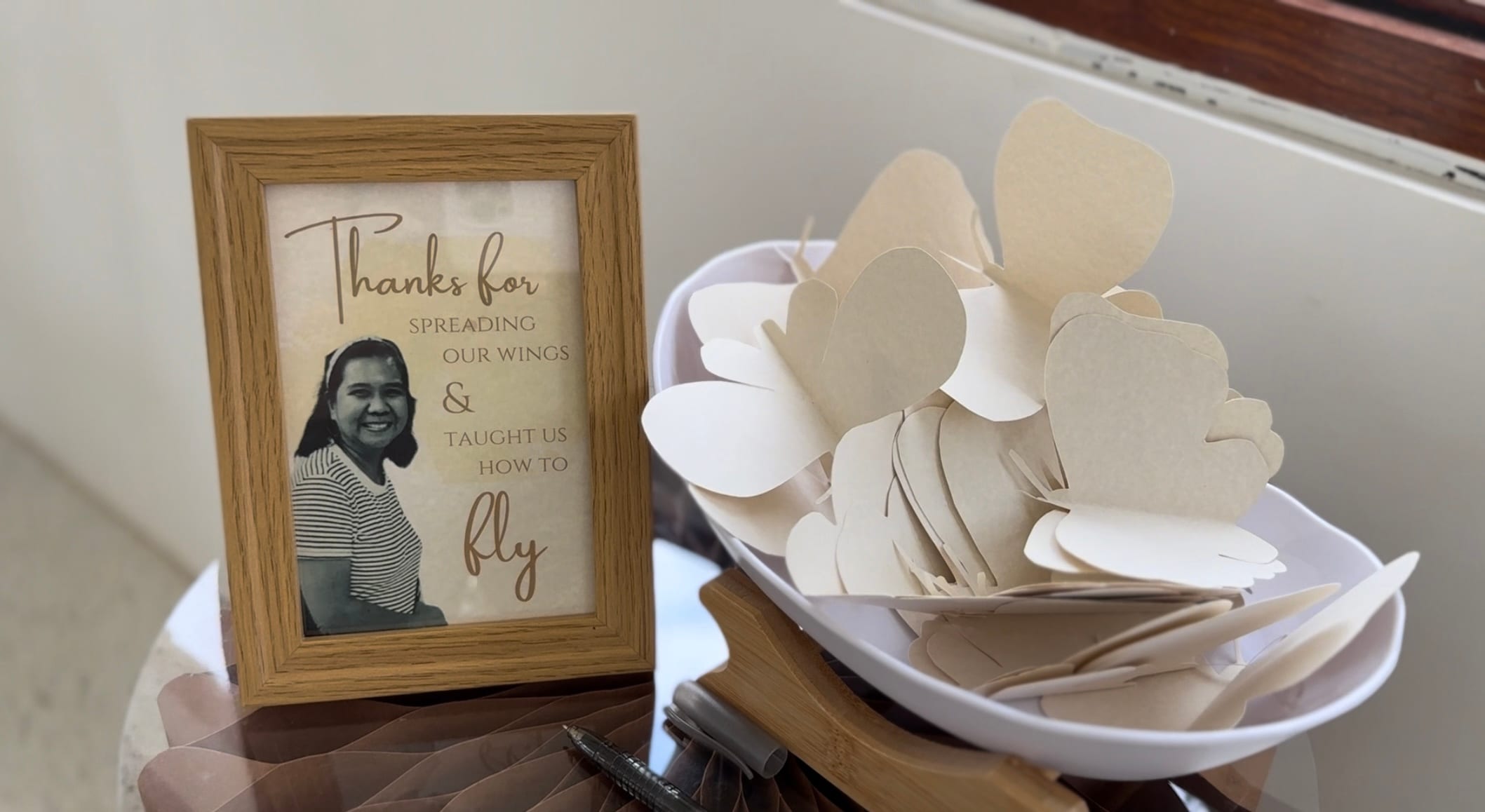 A memorial table with a wooden photo frame tribute and a bowl of wooden butterfly cut-outs for guests to write messages.